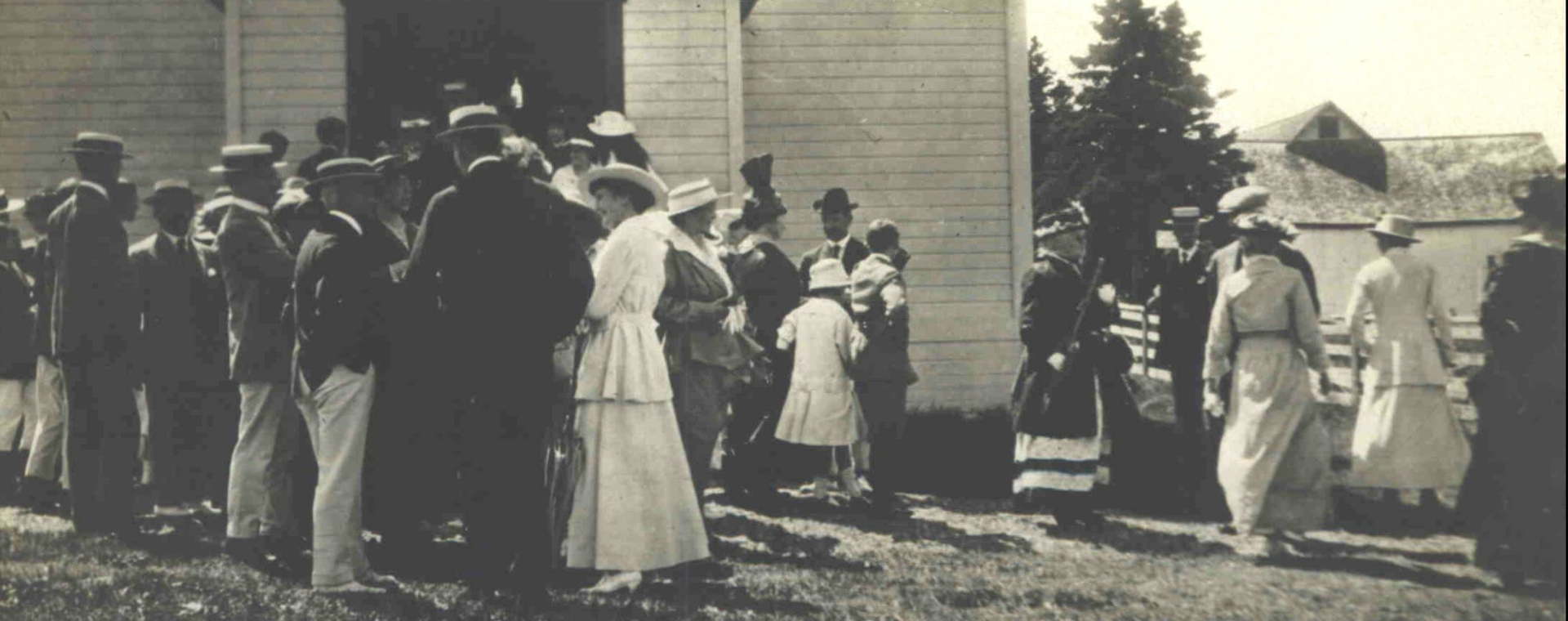 Black and white photograph of men and women gathered in front of a wooden church at the end of mass. The men are, for the most part, dressed in a dark jacket, a white pentalon and a flat braided hat decorated with a ribbon. The women wear a long white dress and a flat braided hat. In the background, a farm building can be seen behind a clump of mature spruce trees.