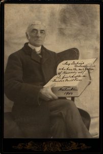 Sepia portrait: seated elderly man, light gray hair, dark suit holding an open book on his lap. Right page of the book: handwritten inscription