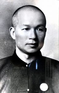 Black and white head and shoulders portrait photograph of a youngish Chinese man with very closely cropped hair, clean shaven, wearing a dark colored Chinese-style collarless jacket.