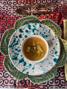 Overhead shot of an orange soup bowl garnished with small diced vegetables, resting on a white decorative plate speckled with blue dots, which is itself placed on a large green leaf-shaped plate. The ensemble is set on a patterned red and blue tablecloth.