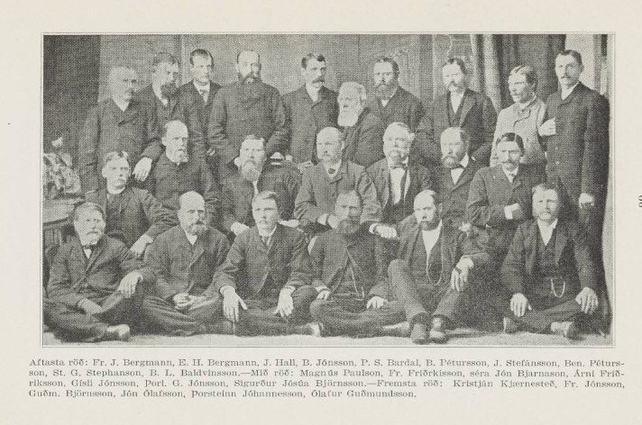 Black-and-white photograph of a group of 30 men posed in three rows, wearing formal 19th-century suits. Some men are seated cross-legged on the floor in the front row, others are seated on chairs in the middle, and the back row stands. Most have mustaches or beards, and their expressions are serious. Below the image is a caption in Icelandic listing the names of all the individuals in the photograph, including Fr. J. Bergmann, St. G. Stephanson, and Ólafur Guðmundsson. The background features a plain curtain.