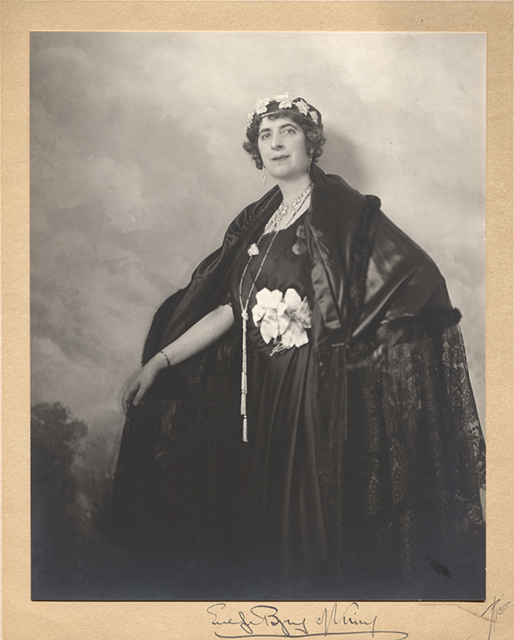 Black and white studio portrait of Lady Byng. Lady Byng poses in front of a painted background of clouds and trees. She is wearing a black cape of satin, lace and fur. A black dress and two white flowers in fabric at the belt. She wears a crown made of maple leaves. Her left hand is resting on her hip allowing to see the details of her cape and dress. Her right arm is outstretched and she is holding her cloak to leave it open. The photograph is mounted on a card signed by her hand
