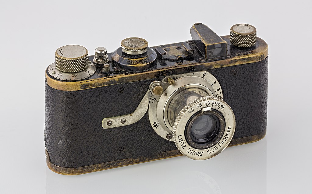 A handheld camera with a leather body and metal lens assembly. On top of the camera are various knurled metal knobs and buttons.