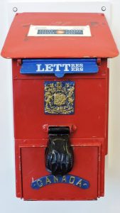 An old style red metal mailbox with a more modern Canada Post logo on top. On the front is a blue flap labeled Letters in English and Lettres in French. Below the flap is an ornate engraving of a Canada’s coat of arms, a black lock cover, and the lettering: Canada.